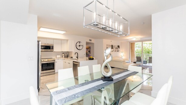 A modern kitchen with stainless steel appliances, a dining area with a glass table and white chairs, and a large window providing natural light.