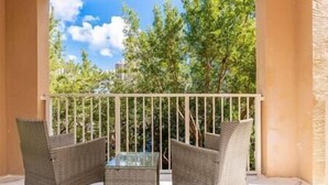 A balcony with wicker furniture and a glass table, overlooking a green area with trees and buildings.