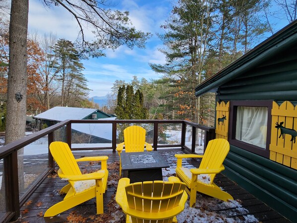 En terrass med gula Adirondack-stolar och ett bord, med utsikt över ett snötäckt landskap med träd och en stuga.