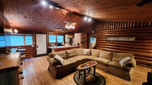 Interior acogedor de una cabaña con techo de madera, un amplio sofá seccional en forma de L, una pequeña mesa redonda y una cocina visible a través de una puerta abierta.