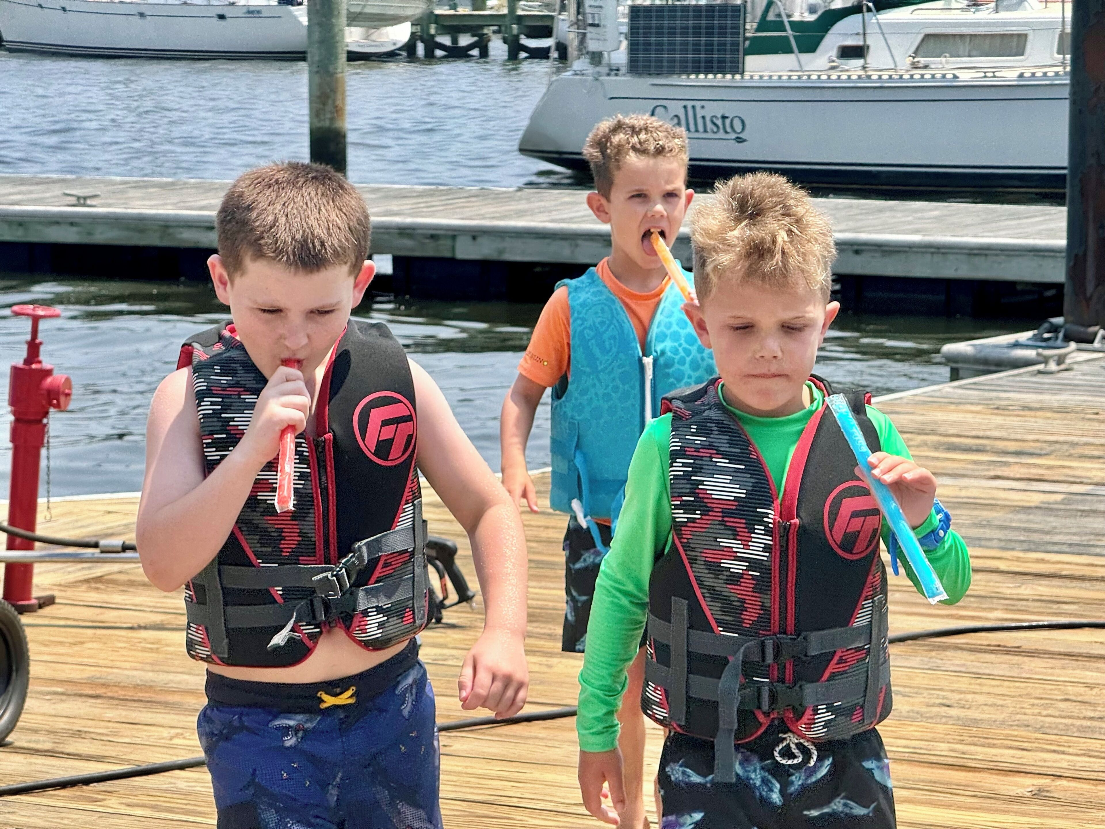 Kinderen met zwemvesten op een steiger, met boten op de achtergrond.