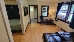 A room with a wooden floor, a black leather sofa, a rocking chair, a small table with a lamp, a TV mounted on the wall, and a window with curtains. - Cabin on 25 Acres of Forest (Bessemer)