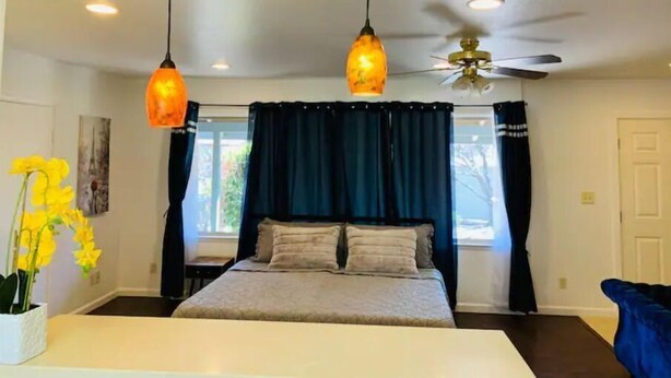 A bedroom with a bed, two pendant lights, a ceiling fan, and a window with curtains. - Full Kitchen Studio w/ AC (Castro Valley)
