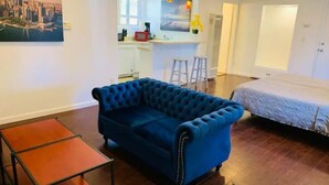 A modern living room with a blue sofa, a small wooden table, a bed with a blue bedspread, and a cityscape painting on the wall. - Full Kitchen Studio w/ AC (Castro Valley)