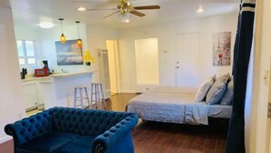 A compact living space with a blue tufted sofa, a bed with a grey duvet, a kitchen area, and a ceiling fan. - Full Kitchen Studio w/ AC (Castro Valley)