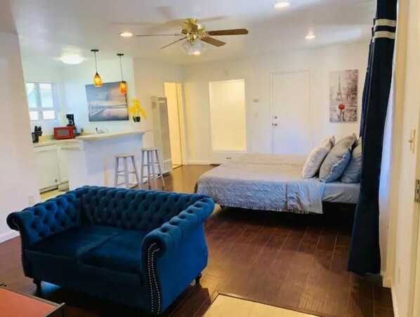 Un espace de vie compact comprenant un canapé bleu à motifs capitonnés, un lit avec une couette grise, un coin cuisine et un ventilateur de plafond.