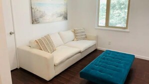 Ein Wohnzimmer mit einem weißen Sofa, einem blauen Hocker, einem Bild an der Wand und einem Fenster mit Blick auf Bäume.