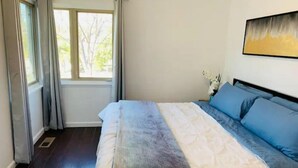 Ein Schlafzimmer mit Bett, einem Fenster mit Vorhängen, einem Nachttisch, einer Vase mit Blumen und einem Bild an der Wand.