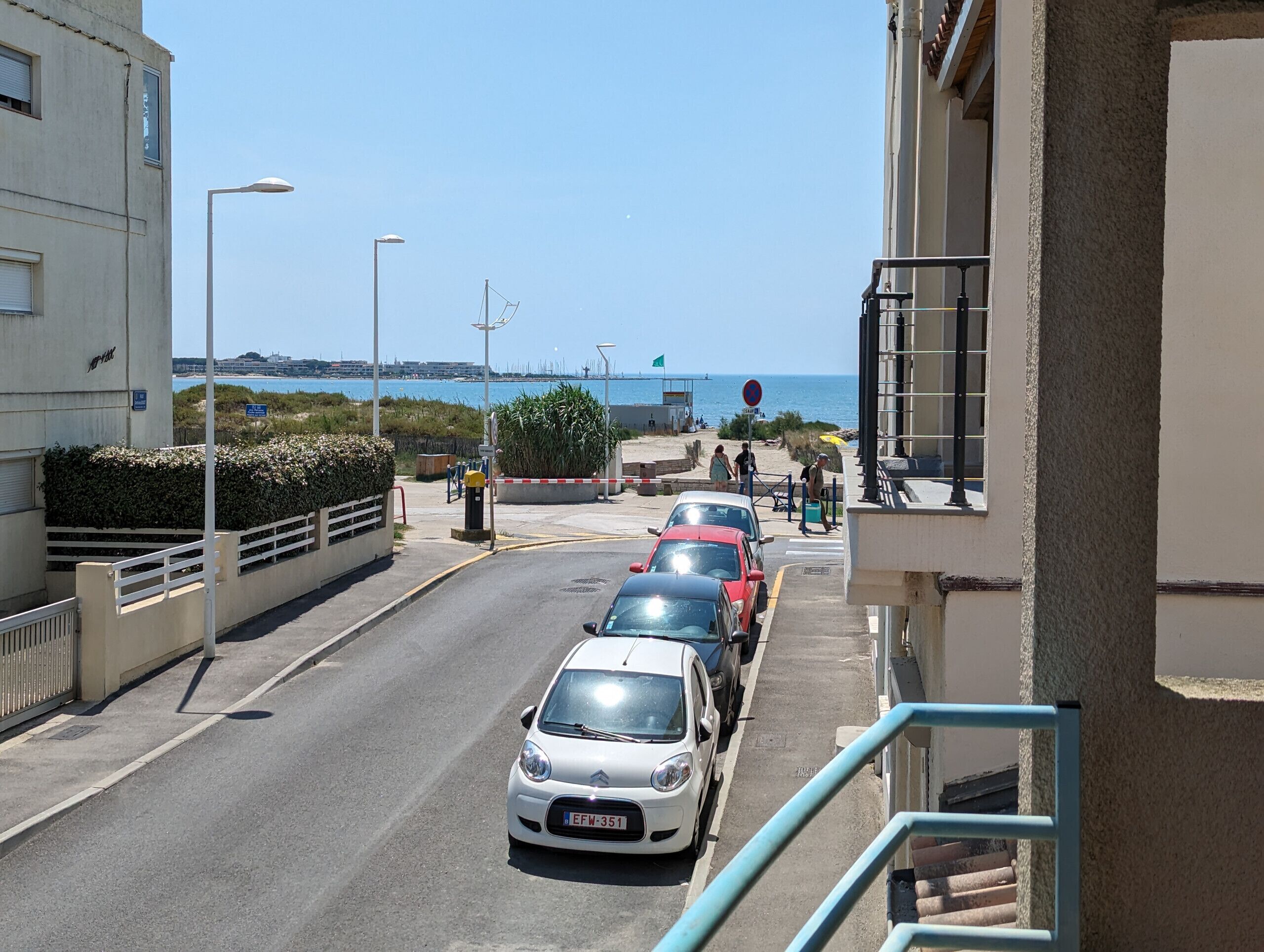 A narrow urban street with parked cars, a pedestrian walkway, and a view of the sea in the background.