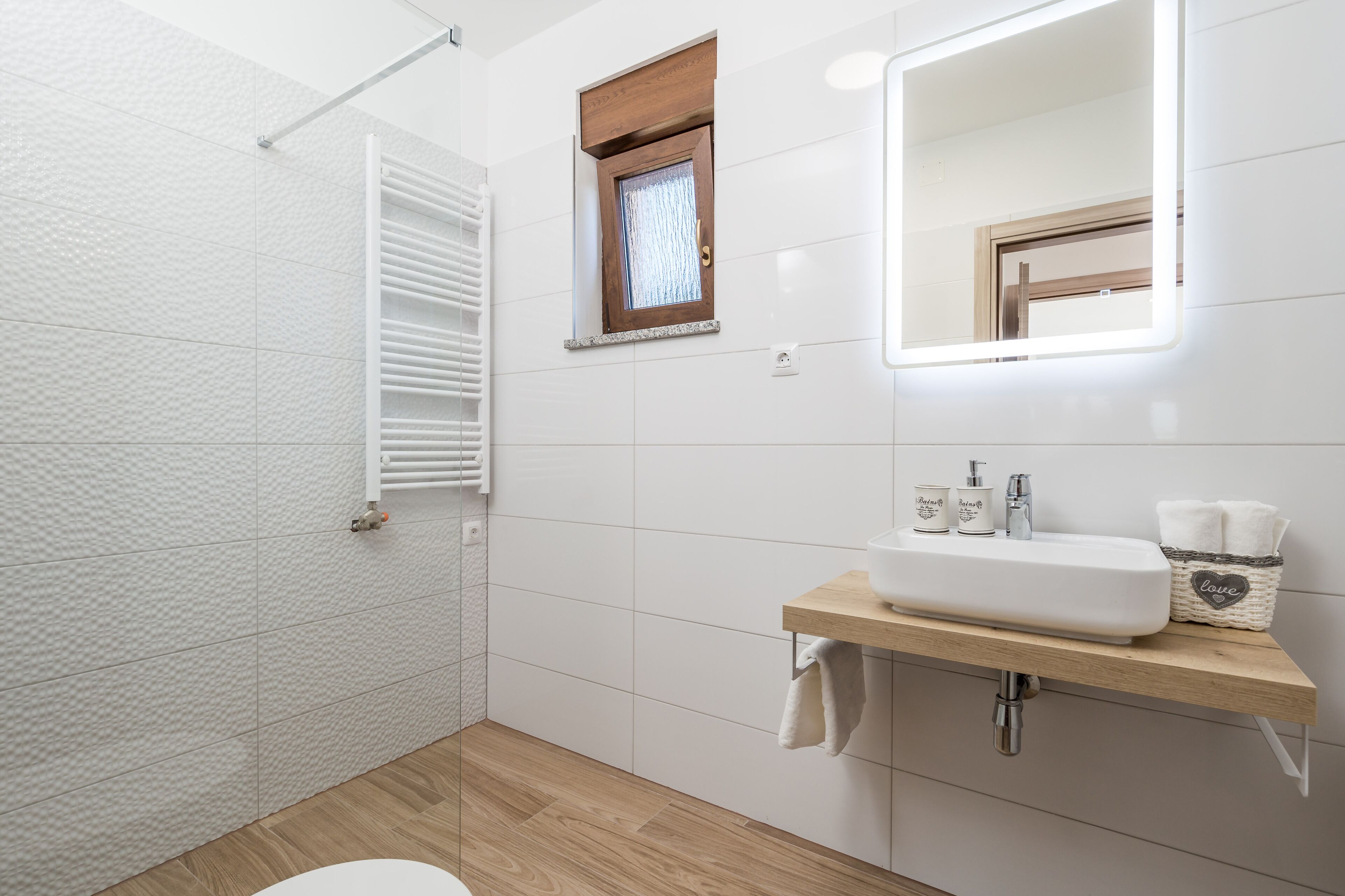 A bathroom with white tiled walls, a wooden floor, a white sink with a wooden countertop, a mirror, and a window with blinds.
