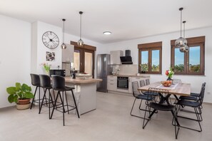 A modern kitchen with a central island, bar stools, a dining table with chairs, a clock on the wall, and large windows.