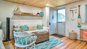 Een gezellige kamer met een houten plafond, een witte bank met turquoise kussens, een kinderstoel, een kleine tafel en een deur die naar buiten leidt.