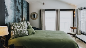 A bedroom with a large bed, a nightstand with a lamp, a chair, a window with curtains, and a painting of a forest on the wall. - Lake Side Guest House with Hot Tub (White Cloud)