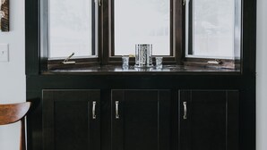 A dark wooden cabinet with three windows above it, a small shelf with a cup and a spoon, and a chair to the left. - Lake Side Guest House with Hot Tub (White Cloud)