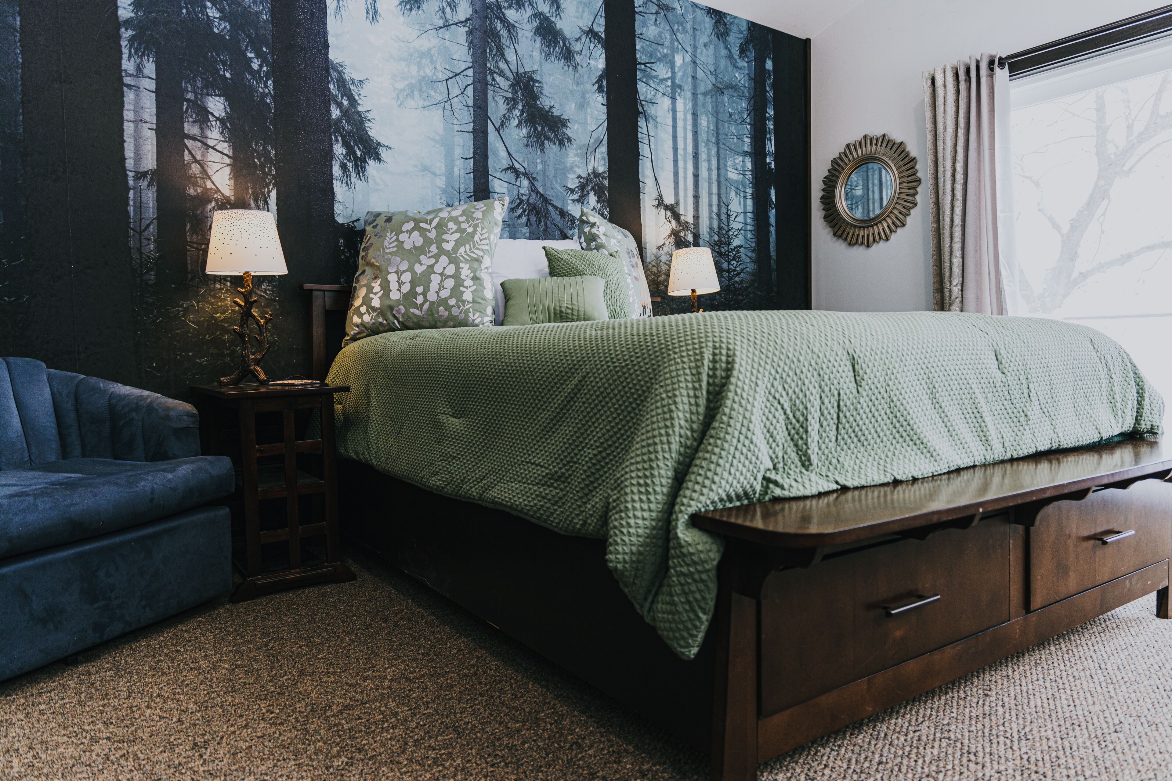 Une chambre à coucher comprenant un lit, deux tables de chevet, une commode, un canapé bleu et une fresque murale sur le thème de la forêt.