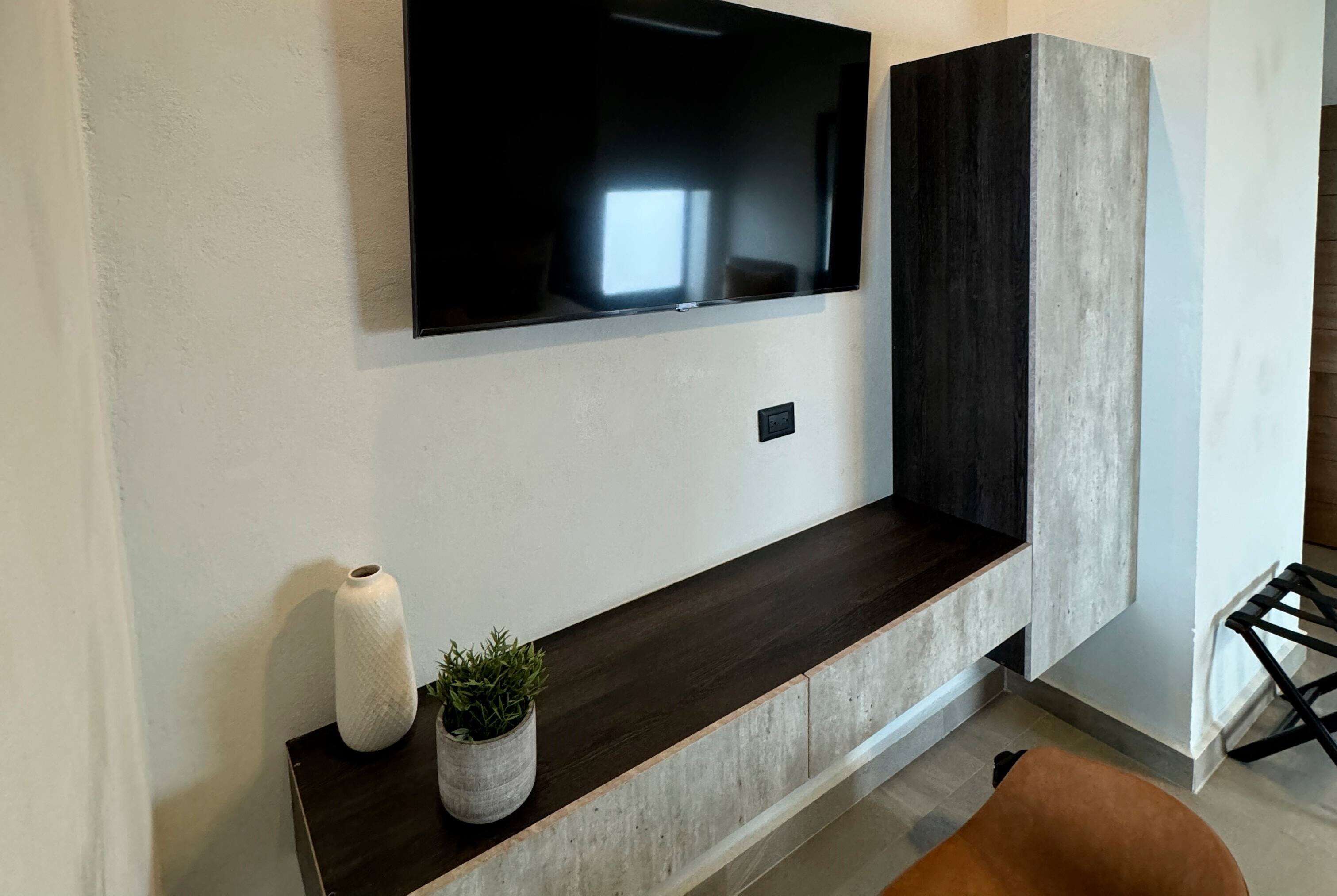 Una sala de estar moderna con un televisor de pantalla plana montado sobre una consola de madera oscura, un jarrón blanco con una planta y un sillón marrón.
