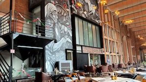 A restaurant with a high ceiling, brick walls, and a mural. Tables are set with chairs and have condiments on them. - Esplendor by Wyndham San Luis Potosi (San Luis Potosi)