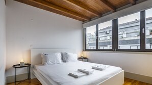 Une chambre moderne avec un grand lit, un plancher en bois, du linge de lit blanc et une vue sur les bâtiments par une grande fenêtre.