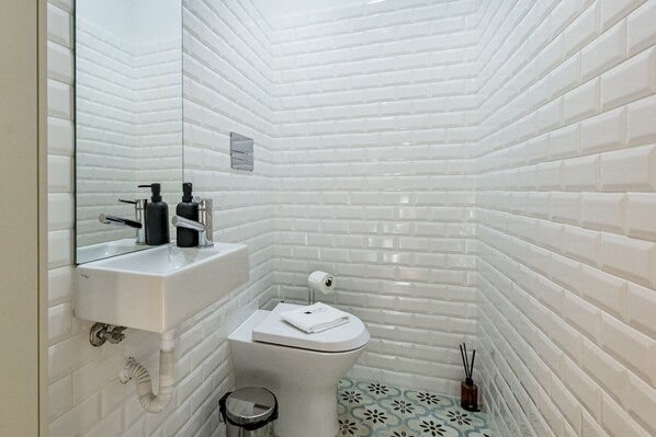A bathroom with white tiled walls, a white sink with a mirror, a toilet, and a small trash bin.