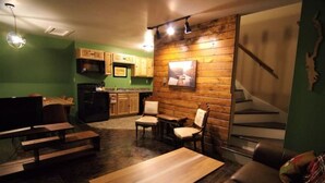 Een gezellige woonkamer met een bank, houten tafels en stoelen, een keuken met houten kastjes, en een trap.