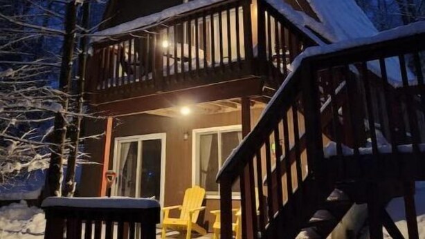 Een houten chalet met een balkon, omringd door besneeuwde grond en bomen, 's nachts.