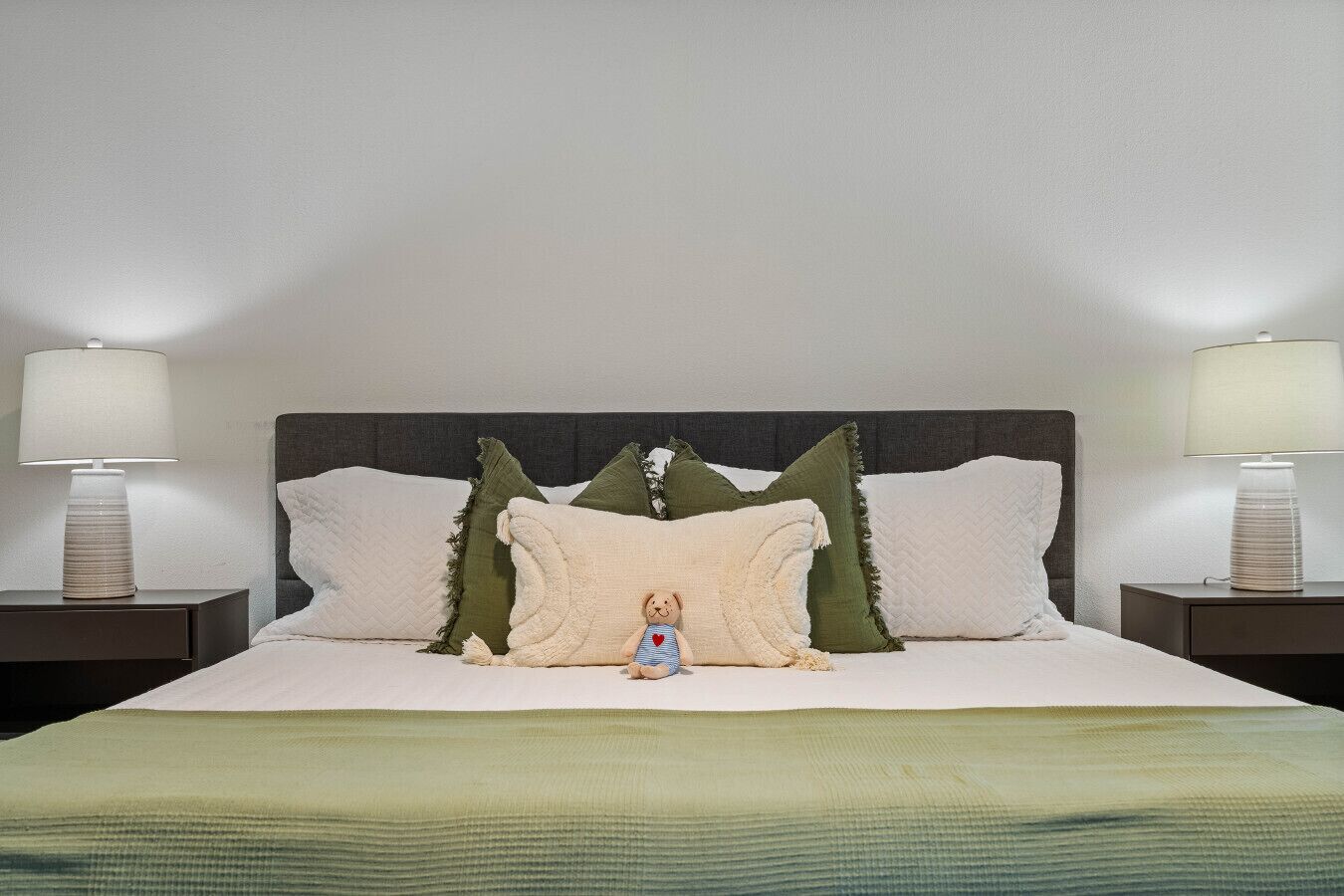 Una cama bien tendida con un edredón verde, dos mesitas de noche con lámparas y un osito de peluche sobre la cama.