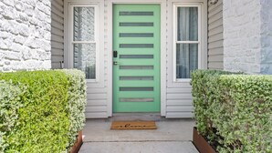 A green door with horizontal slats, flanked by two windows with white frames and curtains, and a welcome mat at the entrance. - Your home away from home! (Round Rock)