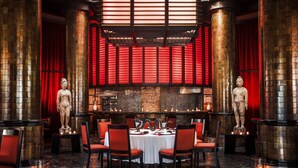 Ein Restaurant mit einem zentralen Esstisch, umgeben von roten Stühlen, flankiert von zwei großen Säulen mit Statuen und einem rot beleuchteten Hintergrund.