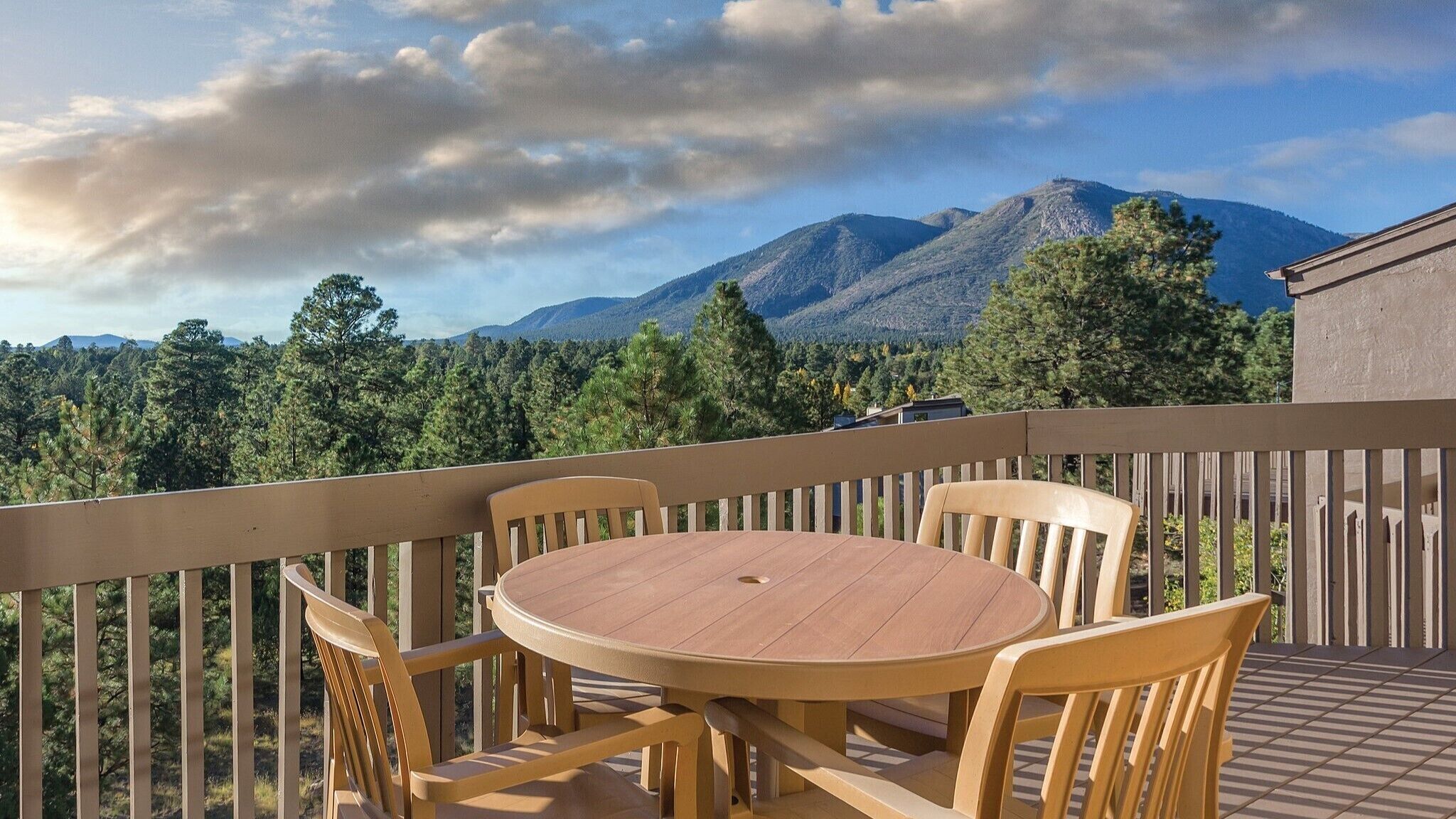 Een houten terras met een ronde tafel en stoelen, met uitzicht op een bergachtig landschap met bomen.