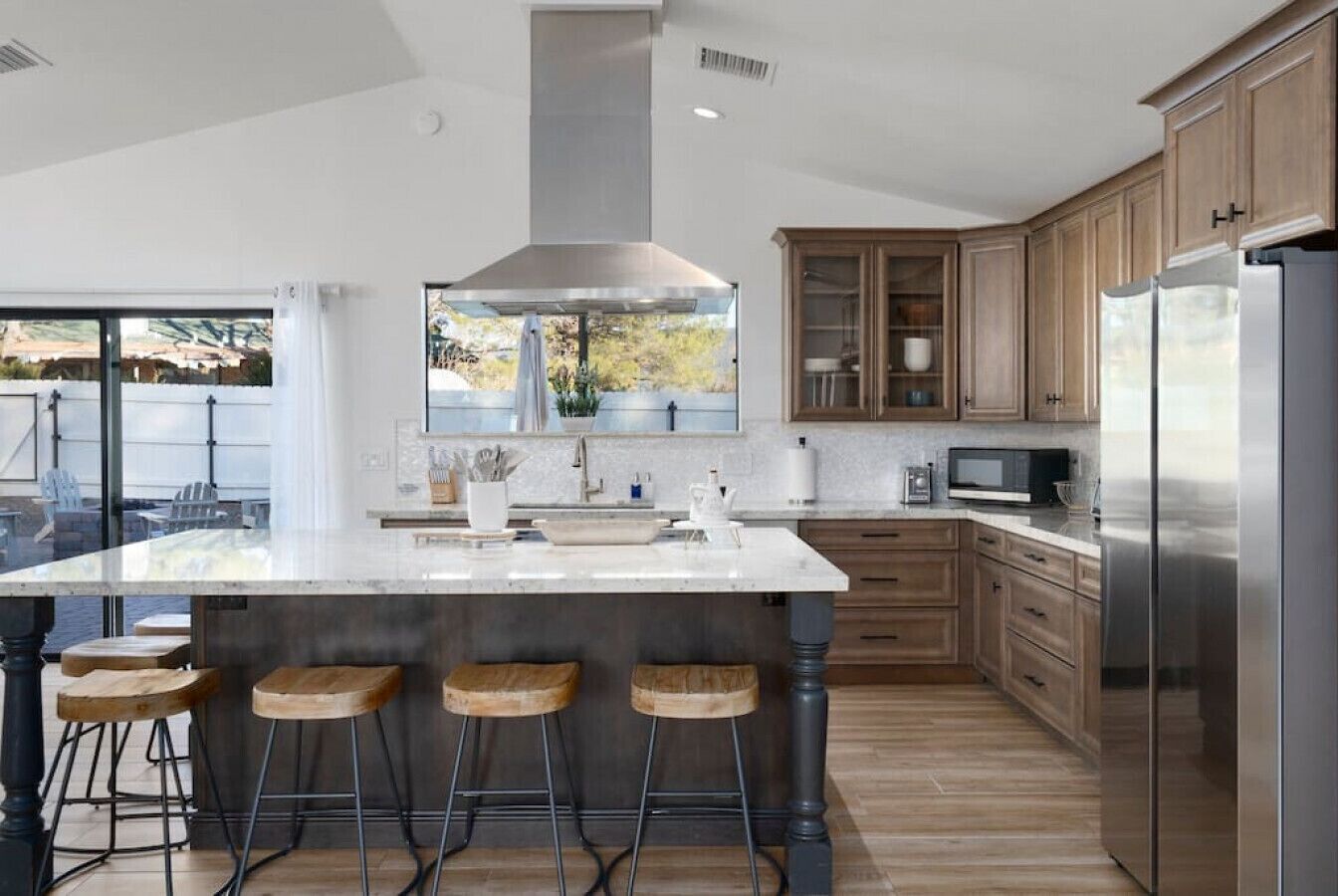 A modern kitchen with a large island, wooden bar stools, stainless steel appliances, and a view through the window.