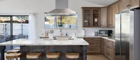 A modern kitchen with a large island, wooden bar stools, stainless steel appliances, and a view through the window.