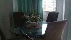 En matplats med ett glasbord, fyra stolar, en krukväxt och utsikt över byggnader genom ett fönster med gardiner.