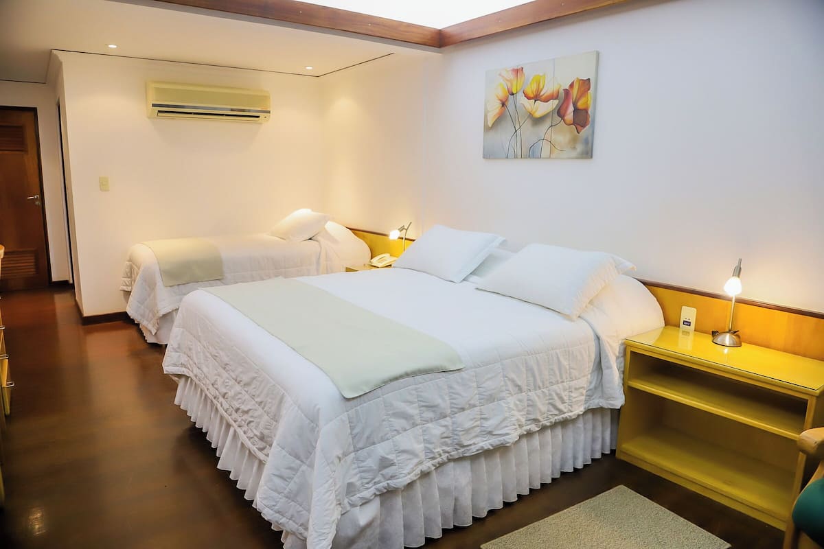 Habitación de hotel con dos camas, una mesita de noche con lámpara, aire acondicionado y un cuadro en la pared.