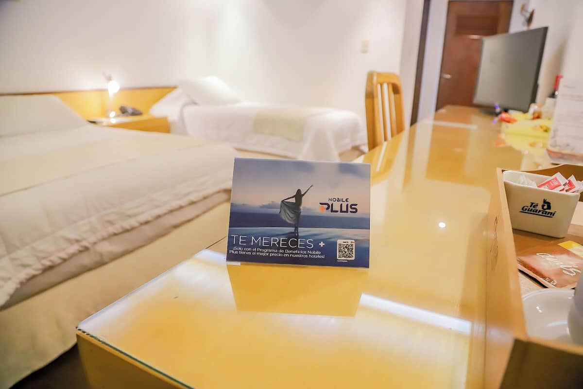 Una habitación de hotel con cama, mesita de noche, televisor y un cartel que anuncia un programa móvil.