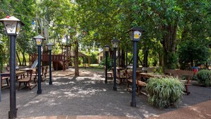 Un área al aire libre con mesas y bancos, rodeada de árboles y un parque infantil.