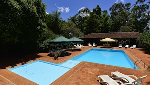 Una piscina con dos zonas separadas, rodeada de sillas de descanso y sombrillas, con árboles y un cielo despejado de fondo.