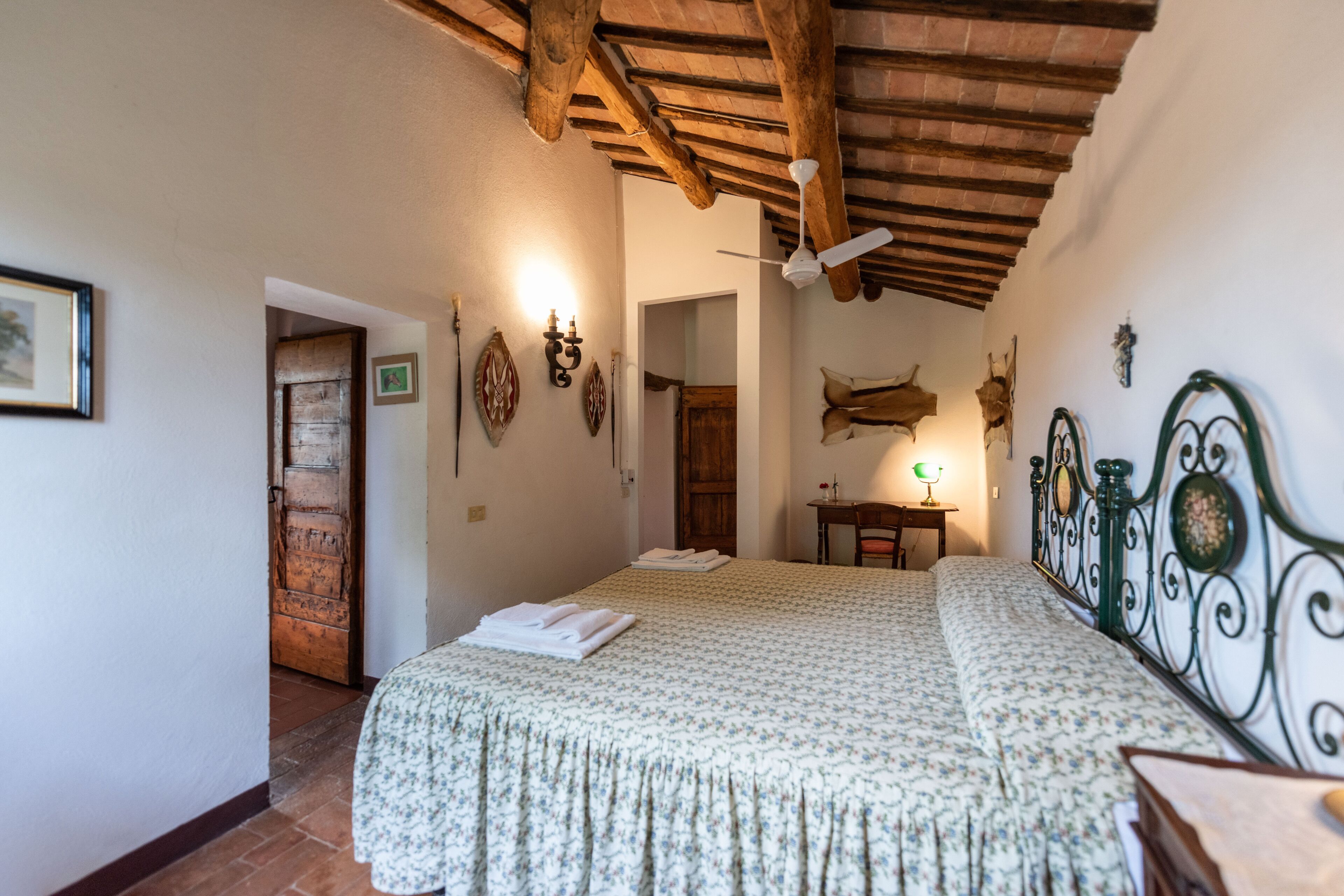 A bedroom with a bed, wooden ceiling beams, a ceiling fan, and a small table with a lamp.