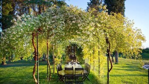 Un área de comedor al aire libre con una mesa dispuesta para cuatro personas, bajo una pérgola adornada con enredaderas florecientes.