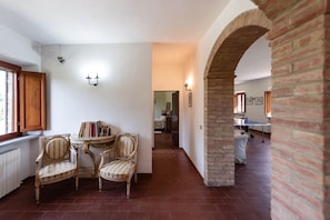 A room with a brick archway, a dining table with chairs, and a view into another room with a bed and a table.