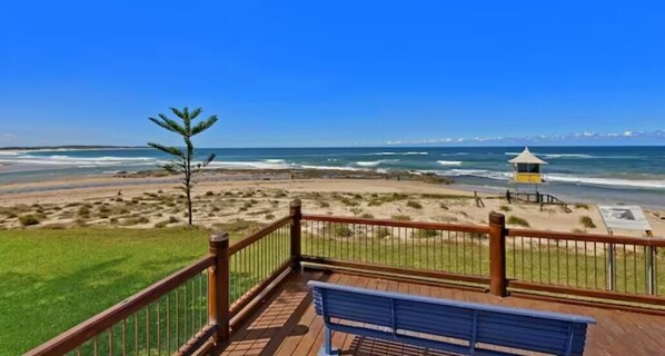 A beach view from a wooden deck with a bench, a lifeguard tower, and a grassy area.