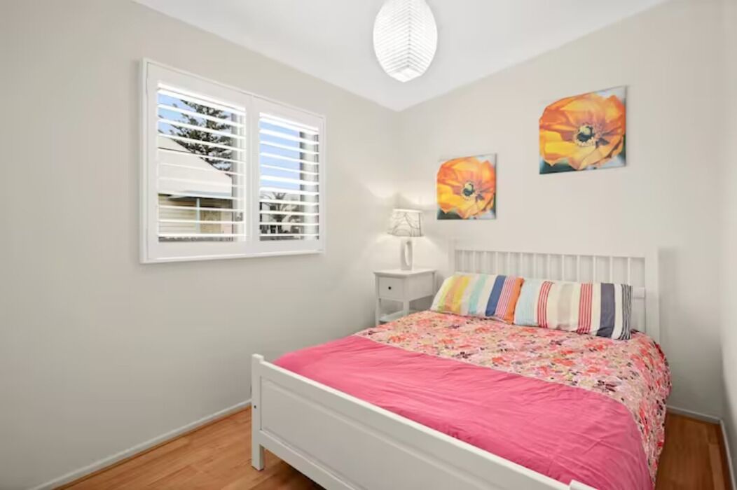 Un dormitorio con cama, una ventana con persianas, una mesita de noche y dos cuadros de flores colgados en la pared.