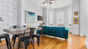 A modern living room with a wooden floor, a teal sofa, a dining table with chairs, a ceiling fan, and a large abstract painting on the wall. - Historic Somerville Cambridge 1 Bed, Inman Square (Somerville)