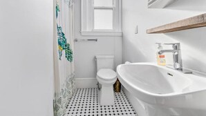 A bathroom with a white toilet, a white sink with a wooden shelf, and a window above the toilet. - Historic Somerville Cambridge 1 Bed, Inman Square (Somerville)