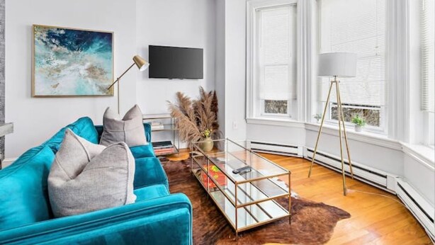 A modern living room with a teal sofa, a glass coffee table, a mounted TV, and a large window with blinds. - Historic Somerville Cambridge 1 Bed, Inman Square (Somerville)