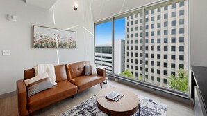 A modern living room with a leather sofa, a round coffee table, and a large window offering a view of a cityscape. - 175 LEX - Walkable Downtown Lexington Condo (Lexington)