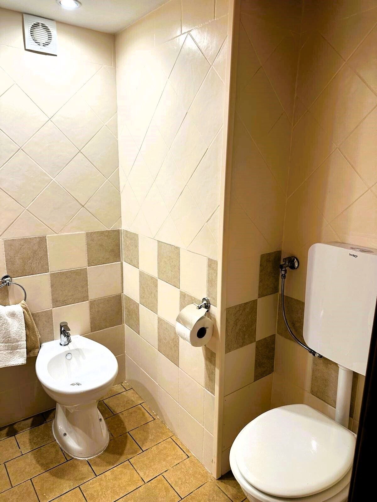 Une salle de bain équipée de toilettes, d’un bidet et d’un lavabo, avec des carreaux de sol aux motifs à carreaux beiges et bruns.