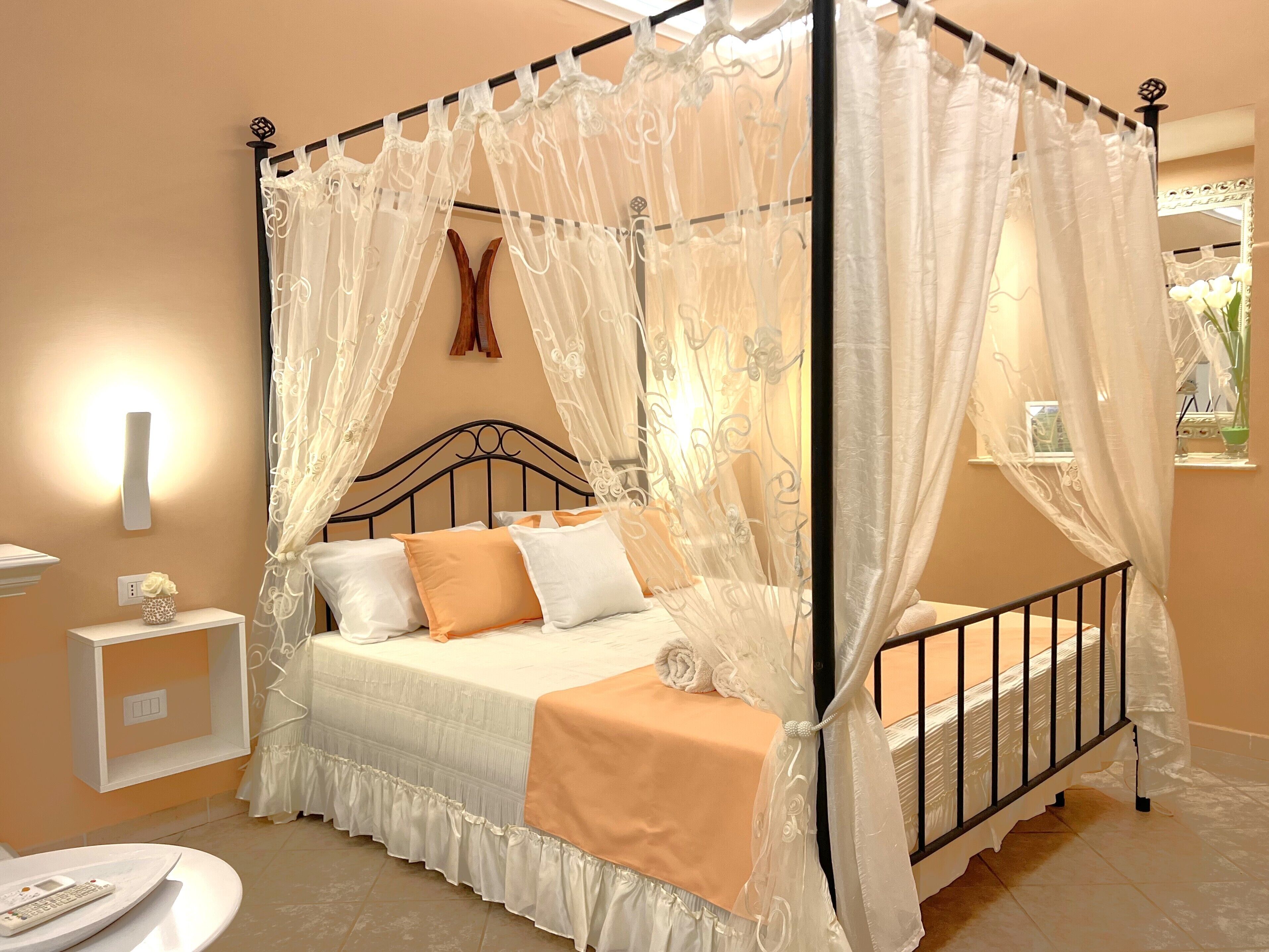 Una cama con dosel y cuatro columnas, con una colcha blanca y almohadas naranjas, una mesita de noche con un florero y una lámpara de pared.