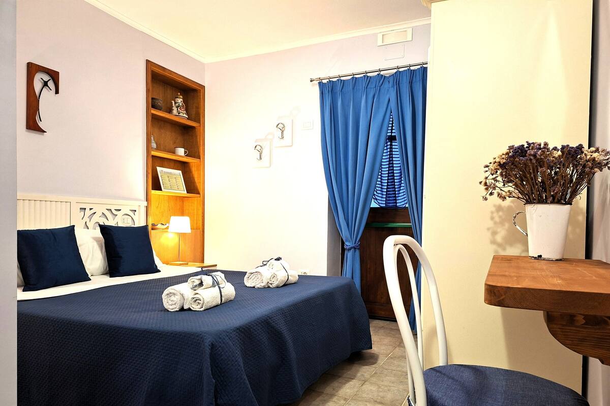 Une chambre d’hôtel comprenant un lit, une chaise, une étagère avec des objets d écoratifs, un rideau et un vase avec des fleurs.