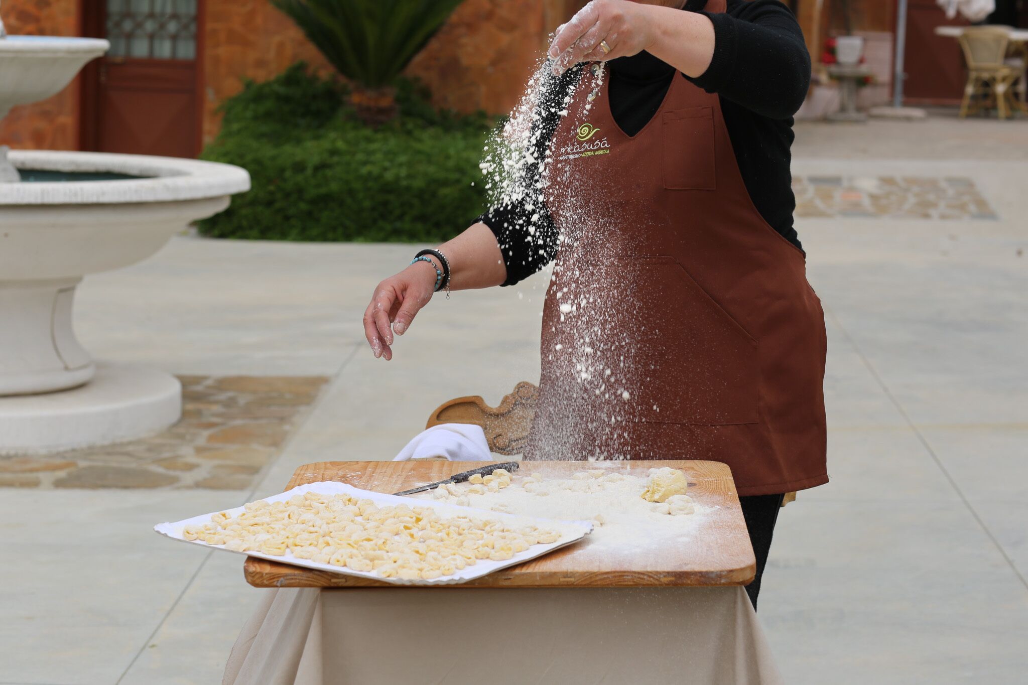 A person in a brown apron sprinkling flour on a wooden table outdoors.