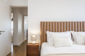 Un dormitorio ordenado con una mesita de noche de madera, una lámpara de noche y una cama con ropa de cama y almohadas blancas.